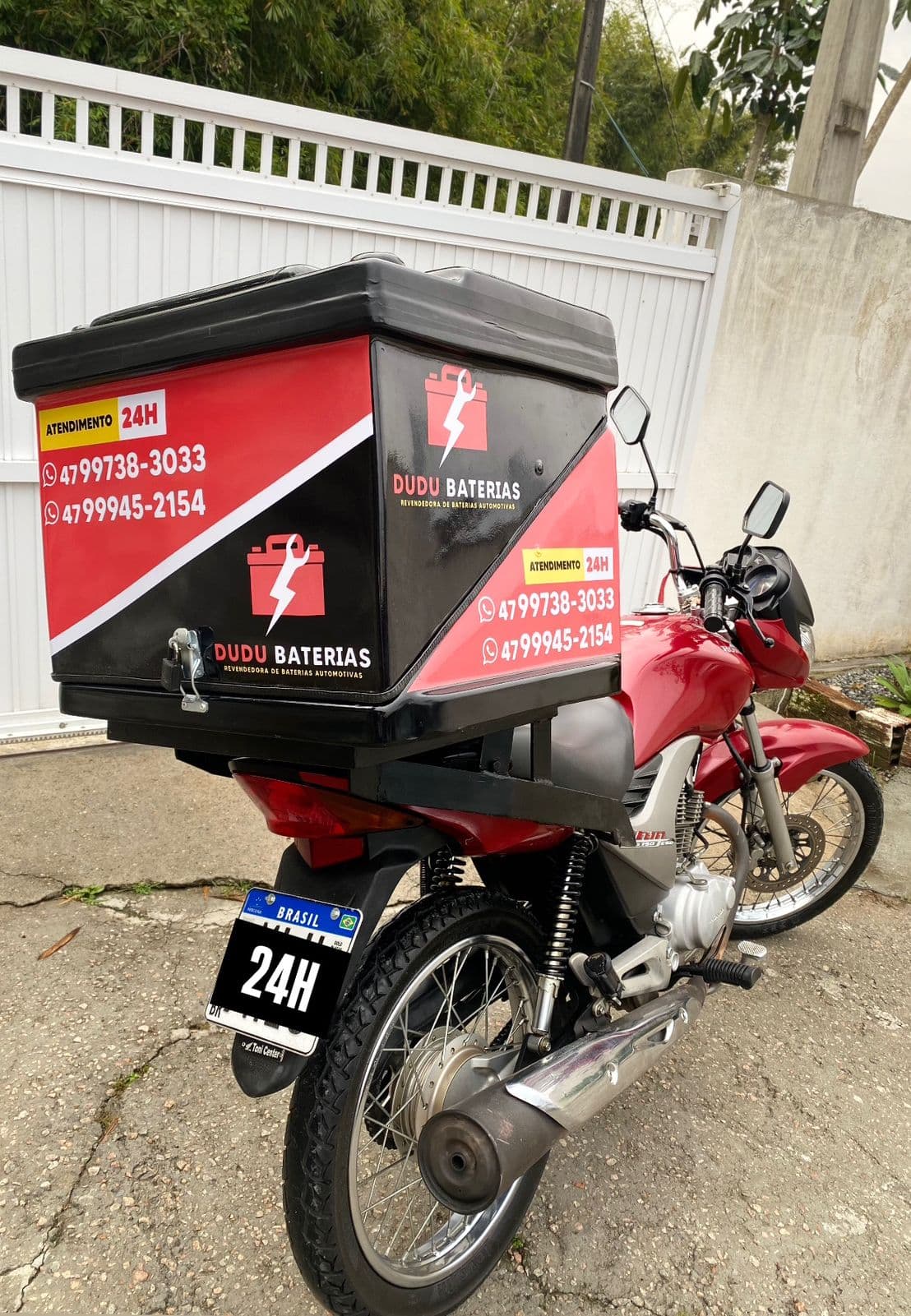 Moto de entrega da Dudu Baterias com baú adesivado e aviso de atendimento 24 horas